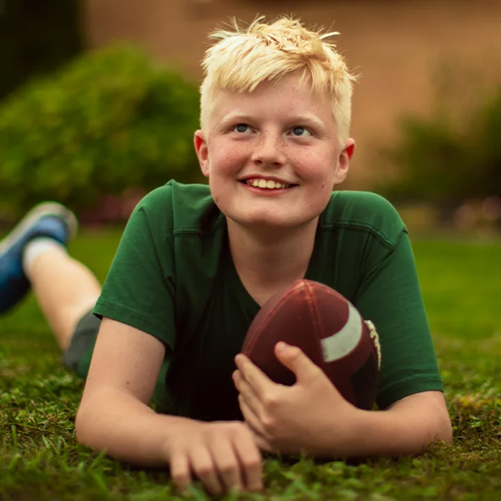 A smiling teenage boy lies in the grass holding a football.